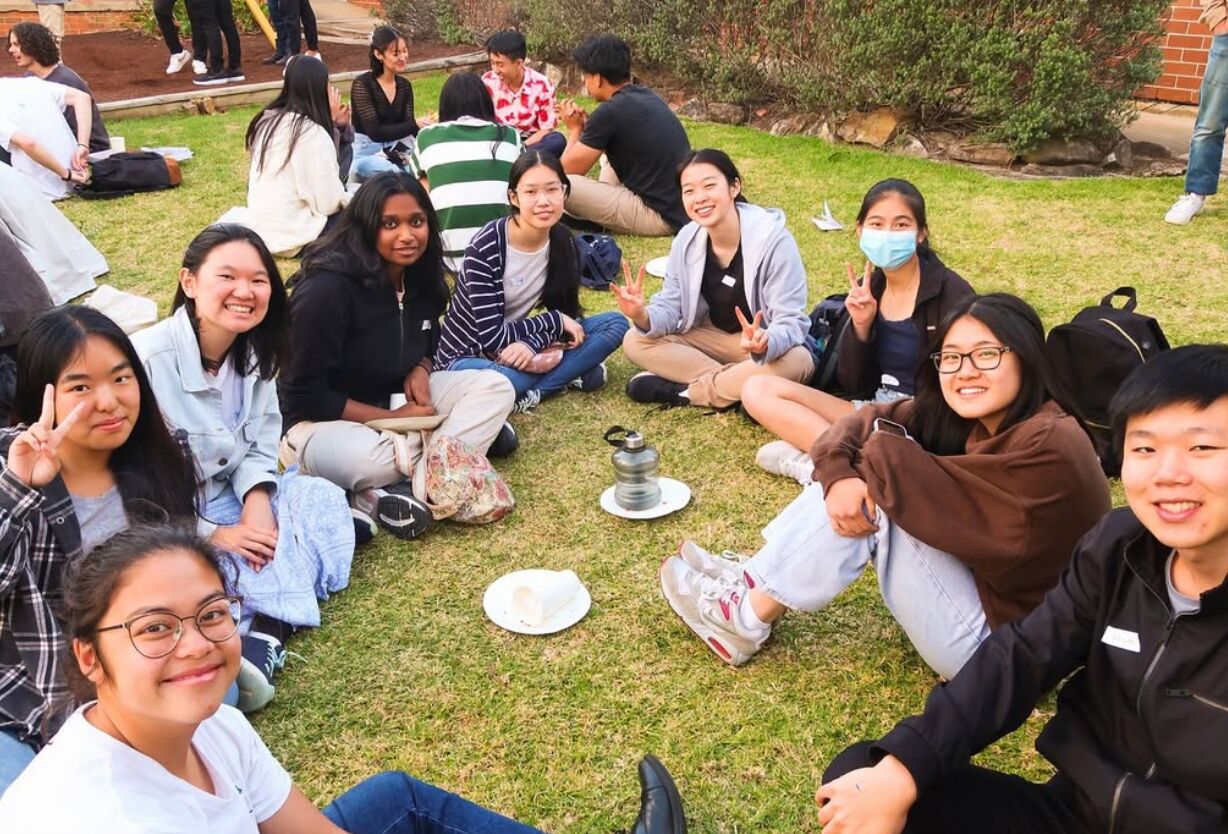 A group of smiling grads sitting together on the lawn, some making peace signs